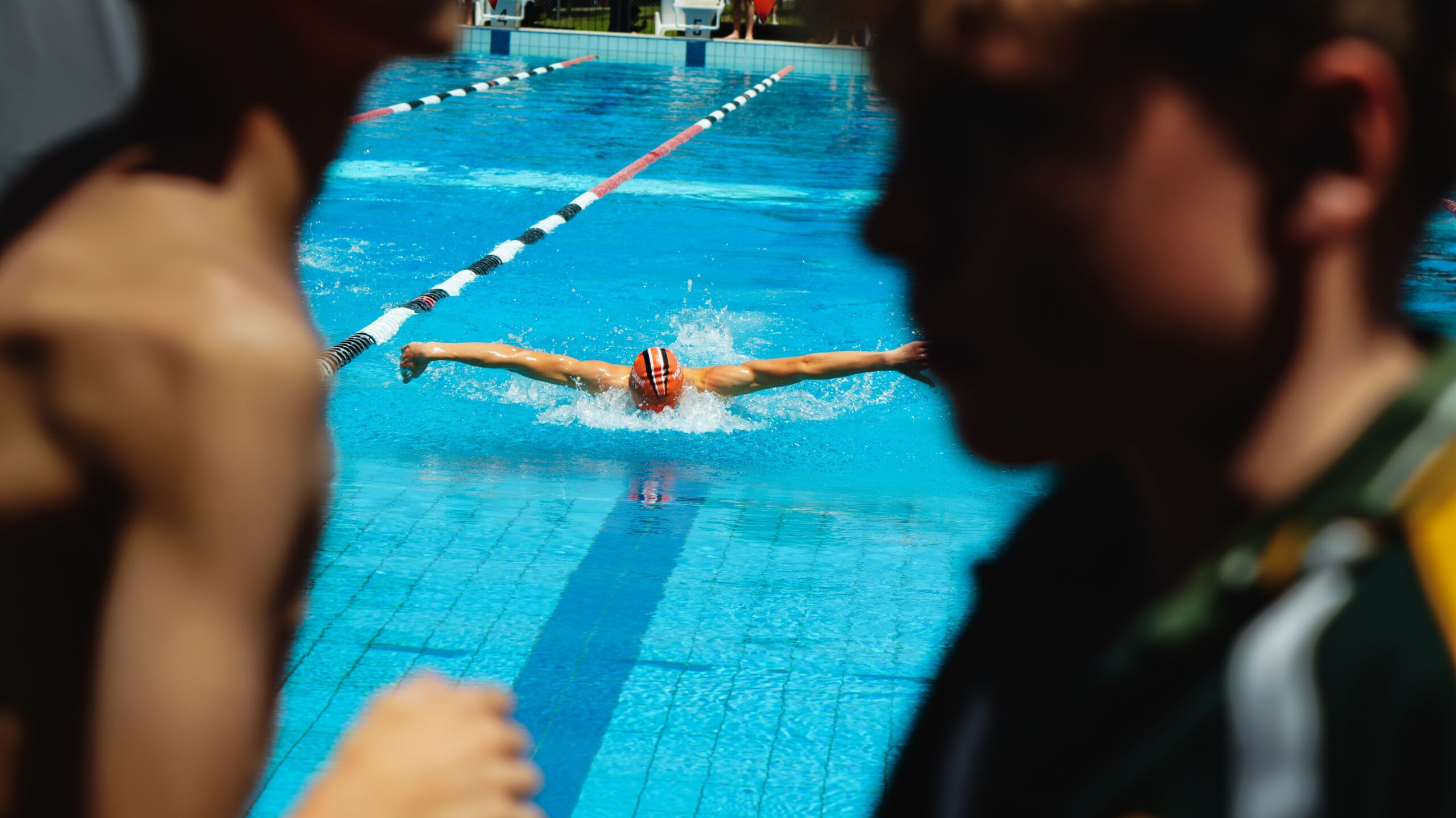 Swimmer performing a butterfly stroke in an outdoor pool with spectators watching.